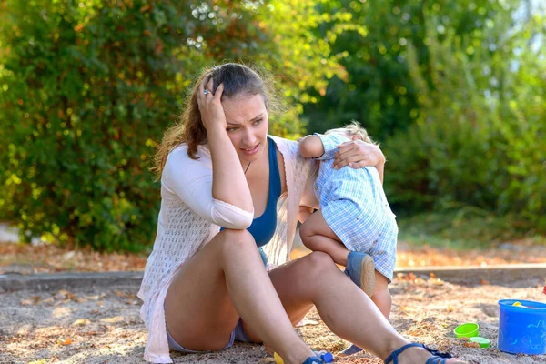 Stressed young mother with a difficult baby boy staring dejectedly at the ground in a playground as the infant acts up as she holds him close to her