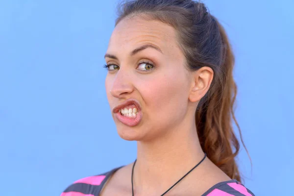 Attractive young woman pulling a funny expression to one side of her face over a blue sky background in a closeup cropped head shot