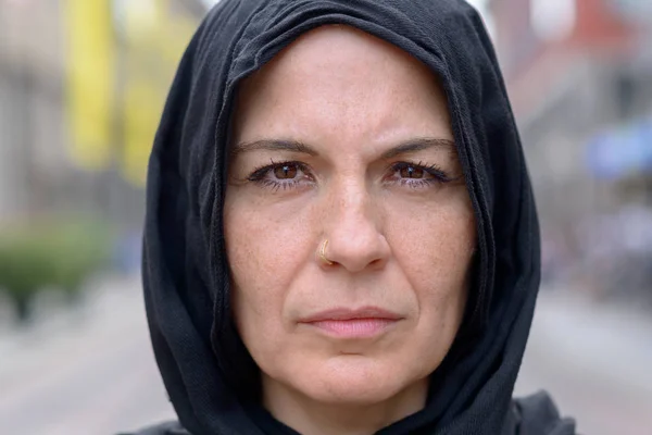 Pensive mature woman wearing a black head scarf staring intently at the camera in a cropped head shot outdoors on an urban street