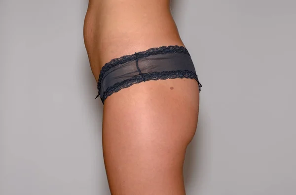 Close up side view of the thigh and hips of a slender fit young woman in black panties over a studio background