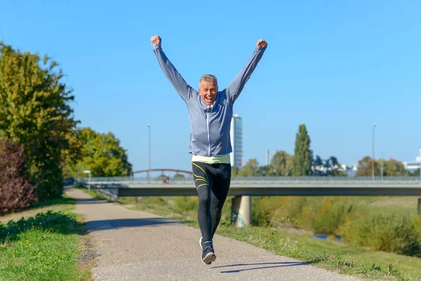 Happy fit middle aged man cheering and celebrating as he walks along a rural lane through a leafy green park after working out jogging