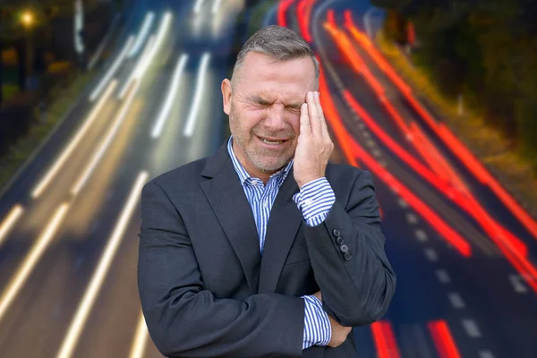 Stressed businessman, or suffering from a headache, standing with his hand to his temple grimacing against Long exposure light trails of traffic on a freeway
