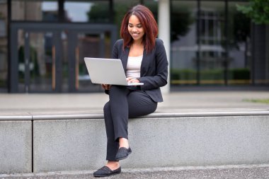 Mutlu bir gülümseme ile bir dizüstü bilgisayar üzerinde çalışan bir ticari bina önünde bir verandada şehirde oturan genç kadın