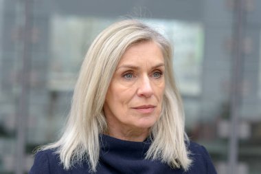 Oldukça düşünceli mavi gözlü sarışın kadın gri bir kentsel arka plan üzerinde rüya gibi uzak bir ifade ile objektif sağa bakıyor