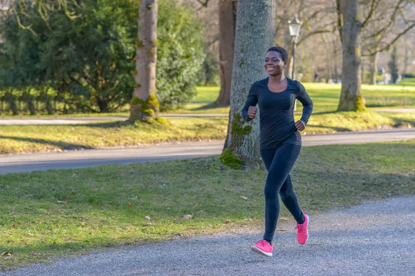 Smiling healthy happy fit young African woman jogging in the park along a tree-lined avenue during her daily exercise workout