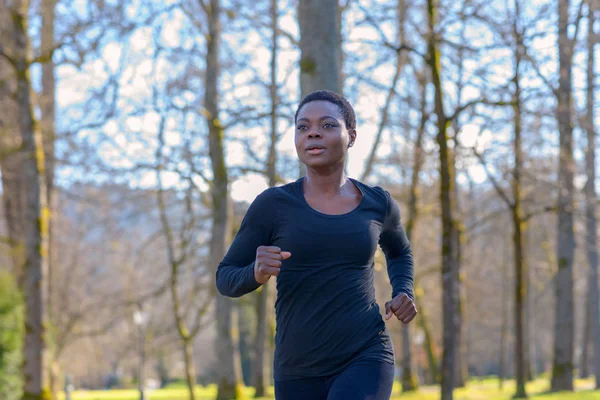 Close up of a determined young African woman training in a park jogging along a tree-lined avenue in a park