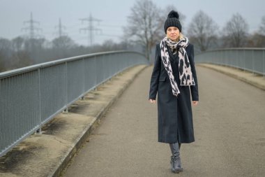 Siyah ceketli ve siyah şapkalı çekici sarışın kadın kameraya doğru yürürken kameraya çok samimi görünüyor.