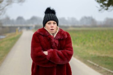 Kırmızı ceketli ve siyah şapkalı çekici sarışın kadın yeşil bir tarlada esniyor.