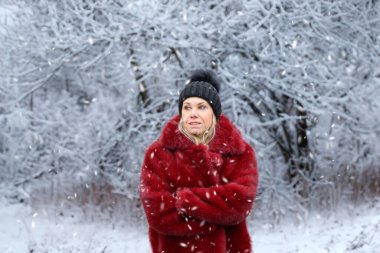 Kırmızı ceketli ve siyah şapkalı çekici sarışın kadın kollarını karlı bir kış manzarasının önünde kavuşturmuş.