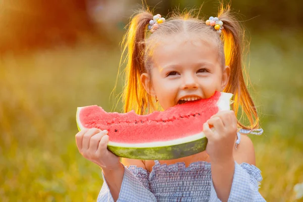 Küçük kız gün batımı ışınları yeşil bir çim üzerinde karpuz bir dilim ısırır
