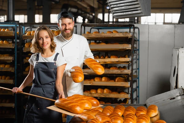 Young bakers with a wooden shovel with bread in their hands against the background of shelving with bread in a bakery. Industrial bread production