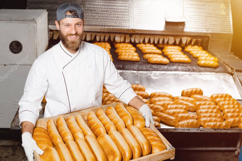 A baker holds a tray with fresh hot bread against the background of an industrial oven in a bakery. Automatic bread making line