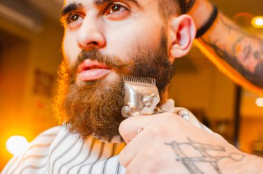 Berber, sakallı ve bıyıklı genç yakışıklı bir adama klasik saç makasından bir sakal kesiyor. Erkekler kuaförü..