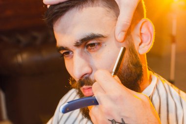 Berber, bıyıklı ve sakallı yakışıklı genç bir adamın tehlikeli tıraş bıçağını kesiyor. Bir erkek kuaförde tehlikeli bir tıraş bıçağı.