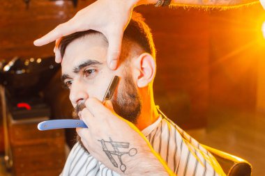 Berber, bıyıklı ve sakallı yakışıklı genç bir adamın tehlikeli tıraş bıçağını kesiyor. Bir erkek kuaförde tehlikeli bir tıraş bıçağı.