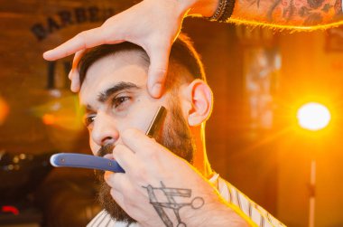 Berber, bıyıklı ve sakallı yakışıklı genç bir adamın tehlikeli tıraş bıçağını kesiyor. Bir erkek kuaförde tehlikeli bir tıraş bıçağı.