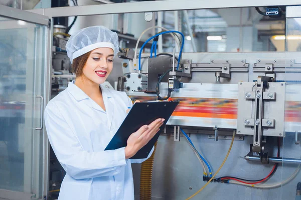 A young beautiful girl in white overalls makes notes in a tablet on the background of equipment of a food processing plant. Quality control in production.