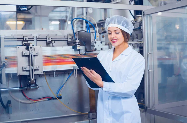 A young beautiful girl in white overalls makes notes in a tablet on the background of equipment of a food processing plant. Quality control in production.