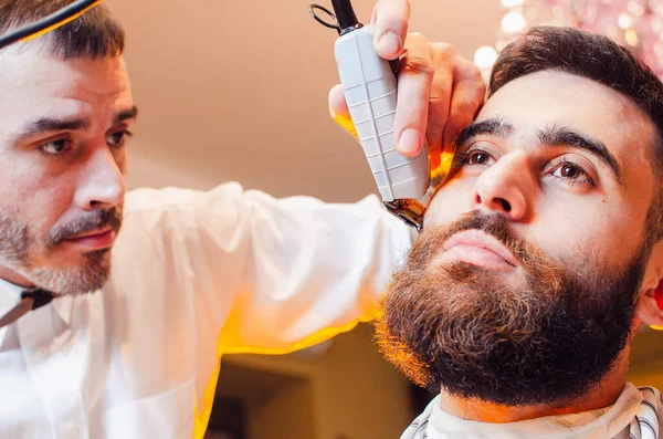 Barber cuts a beard with a trimmer to a young handsome guy with a mustache. Haircut of a beard in a male barber shop.