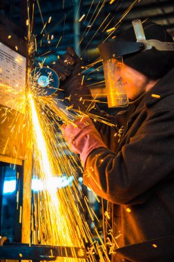 Çalışan ve maske takan bir adam, metalurji sanayi fabrikasının arka planında bir Bulgar ile metal testeresi kesiyor..