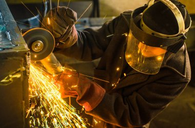 Çalışan ve maske takan bir adam, metalurji sanayi fabrikasının arka planında bir Bulgar ile metal testeresi kesiyor..
