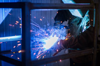 Tulumlu ve koruyucu maskeli işçi kaynak makinesiyle metal kaynaklıyor. Profesyonel kaynakçı. Koruyucu tulumlu bir adam..