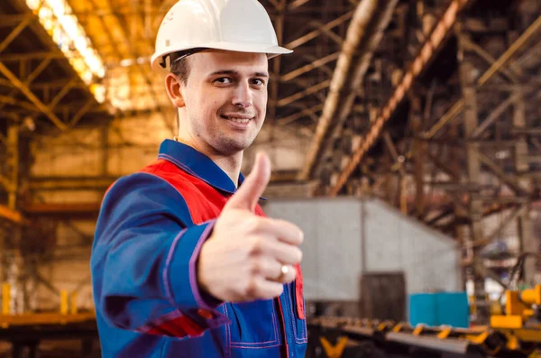 Bir adam baş parmağını gösteriyor. Tulumlu bir çalışan ve endüstriyel bir fabrikanın beyaz kaskı bir sınıfı gösteriyor. Hareketler.