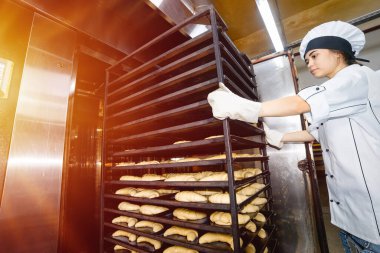 Fırıncı kız, fırındaki endüstriyel bir fırına içi hamur dolu fırın çarşaflarıyla birlikte bir araba yerleştiriyor..