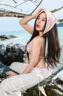 A young woman relaxes at the beach, wearing a colorful bikini and enjoying the sun. The ocean waves and rocky shore create a serene atmosphere.