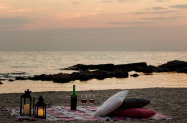 Yaz deniz günbatımı. Sahilde romantik piknik. Bir şişe şarap, Gözlük, mumlar, ekose ve yastıklar. Seçici odak.