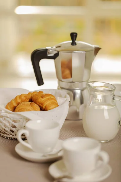 Coffee maker, cups and croissants in the basket are on a round table in front of the window. Selective focus.