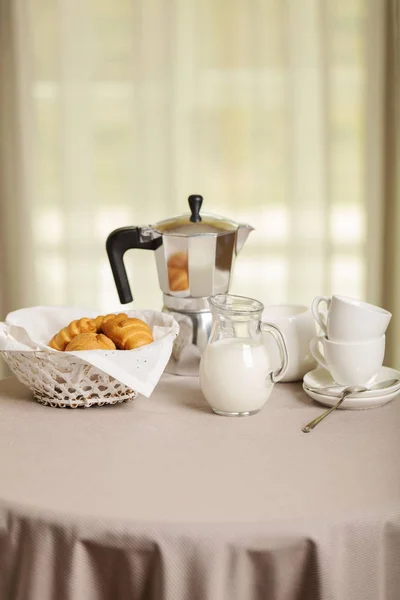 Coffee maker, cups and croissants in the basket are on a round table in front of the window. Selective focus.