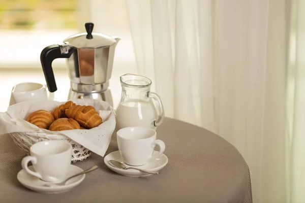 Coffee maker, cups and croissants in the basket are on a round table in front of the window. Selective focus.