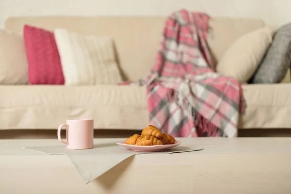 Beige sofa with plaid, colorful pillows (pink, grey, white) and coffee with croissantsin the living room. Selective focus.