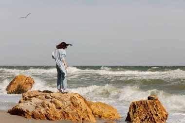 Genç kadın (esmer) bir ışık mavi hırka ve kot pantolon, bir sırt çantası ile sahilde yürüyor. Seçici odak.