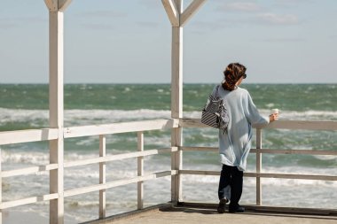 Genç kadın (esmer) bir ışık mavi hırka ve donanma kot pantolon, çizgili bir sırt çantası ile plaj boyunca yürür ve kahve içiyor. Seçici odak.