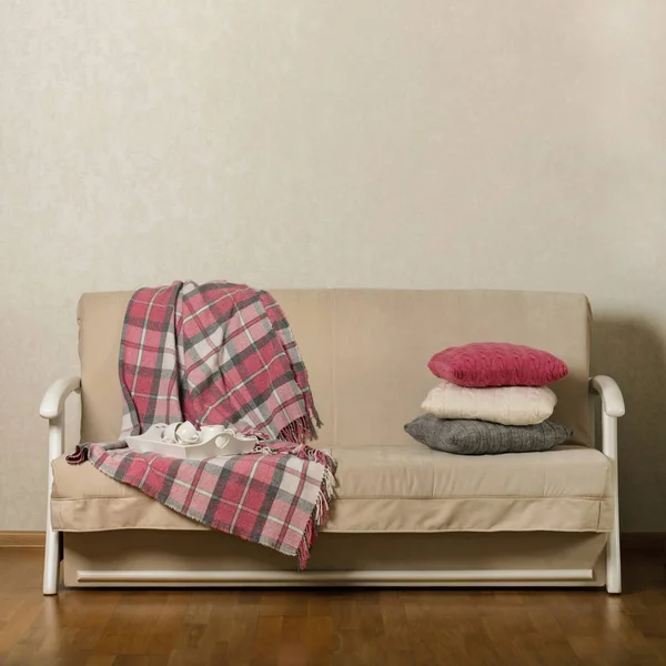 Beige sofa with plaid and colorful pillows (pink, grey, white) and tray with cups of coffee in the living room. Selective focus.