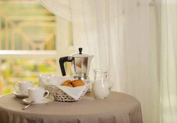 Coffee maker, cups and croissants in the basket are on a round table in front of the window. Selective focus.