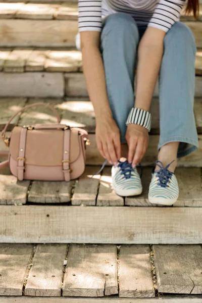 On the back of a blurry background young woman in blue jeans and striped sneakers sits on old wooden steps and ties up shoelaces. Selective focus.