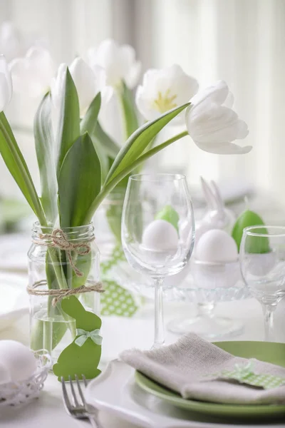 Decor and table setting of the Easter table is a vase with white tulips and dishes of green and white color. Easter decor with white polka dots. Selective focus.