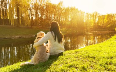 Genç kadın ve gün batımında nehir kıyısında oturan golden retriever köpek