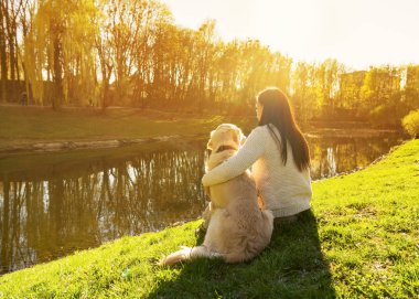 Genç kadın ve gün batımında nehir kıyısında oturan golden retriever köpek
