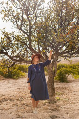 Güzel sarı saçlı genç kadın elma ağacı yakınında mavi etek ve kahverengi şapka giyiyor. Yerel tarım iş büyüme kavramı. Yavaş yaşam otantik yaşam tarzı.