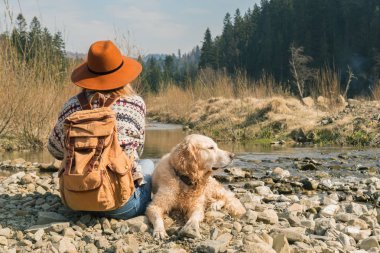 Doğada seyahat eden kadın ve altın retriever köpek. Dağlar nehir ve orman yakınında vintage tekstil sırt çantası ile kahverengi şapka ve yün kazak şık hipster. Seyahat ve wanderlust kavramı. 