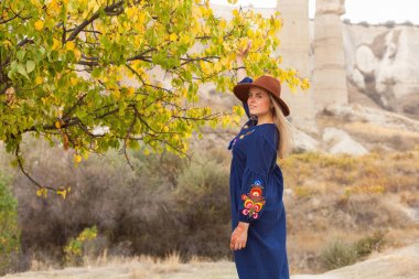 Goreme, Kapadokya Love Vally sonbahar ağacı yakınında otantik mavi etek ve kahverengi şapka giyen güzel sarı saçlı kadın seyahat. Kopya alanı, metin için yer