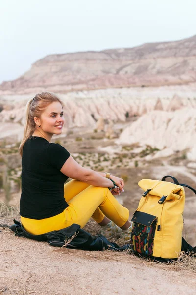 Smiling beautiful blond hair woman with yellow authentic backpack looking on mountains valley. Travel adventures in desert. Goreme, Cappadocia, Turkey.  Copy space background, empty place for text.