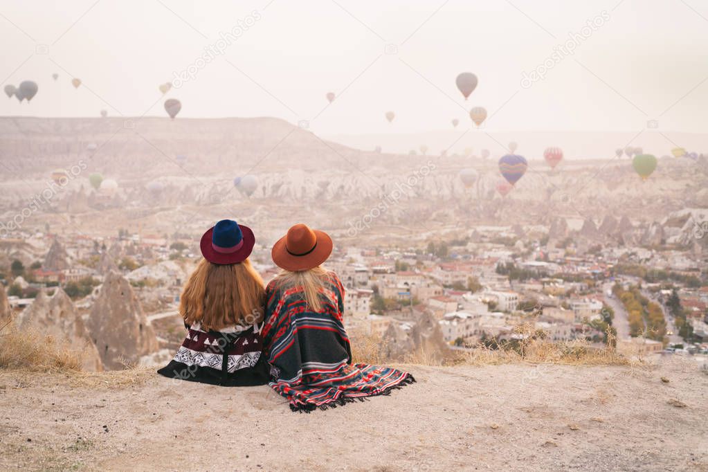 Traveling women wearing authentic boho chic style poncho, sweeter and hats looking on air ballons in sky in Cappadocia valley. Travel and wanderlust concept. Copy space background.