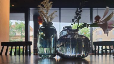 Taze çiçekler içeren üç zarif cam vazo, büyük pencerelerden gelen doğal ışıkla dolu sıcak ve davetkar bir alanda, ahşap bir masanın üzerine yerleştirilir..