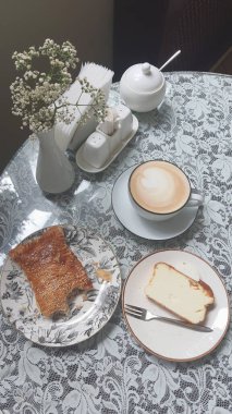 Sıcak bir fincan kahve bir dilim pasta ve dantel kaplı bir masanın üzerinde bir parça hamur işi yanında oturur. Çiçekli küçük bir vazo rahat kafe atmosferine cazibe katar..