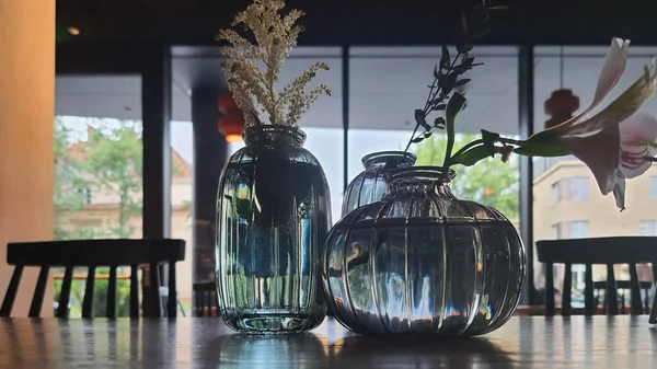 Taze çiçekler içeren üç zarif cam vazo, büyük pencerelerden gelen doğal ışıkla dolu sıcak ve davetkar bir alanda, ahşap bir masanın üzerine yerleştirilir..
